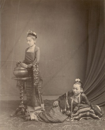 Old photograph from the late 1800s of two wealthy Burmese women. One is standing at a table opening an urn. The other is reclined on a pillow on the floor.
