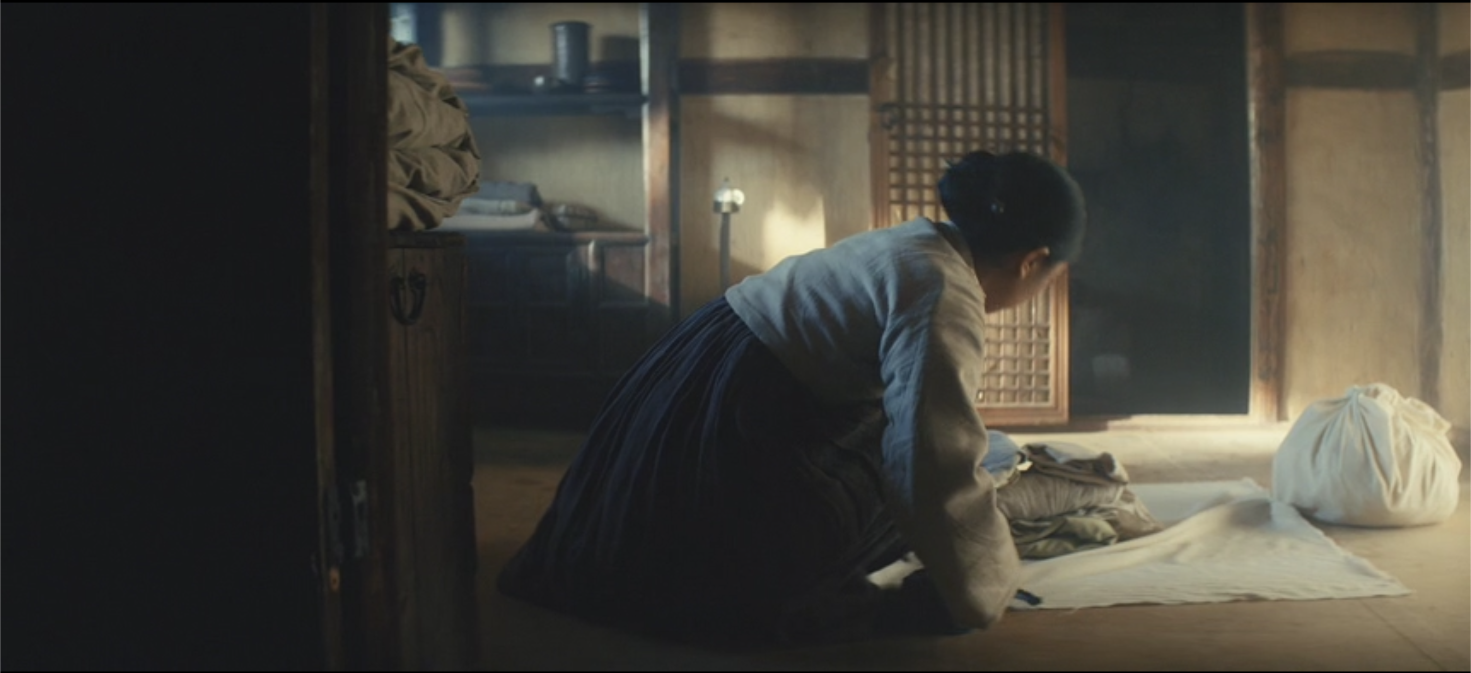 On the 1930s set, Yangjin packs her daughter Sunja's belongings ahead of the latter's journey to Osaka. As the camera slowly tracks left, Sunja's room in Osaka fifty years later appears in the off-screen space to the left. Image capture, Pachinko (2022).